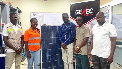 Five people are standing indoors around a large solar panel, with a "GENEC Electrix Ltd." banner behind them. The setting is an office with technical drawings on a whiteboard in the background.